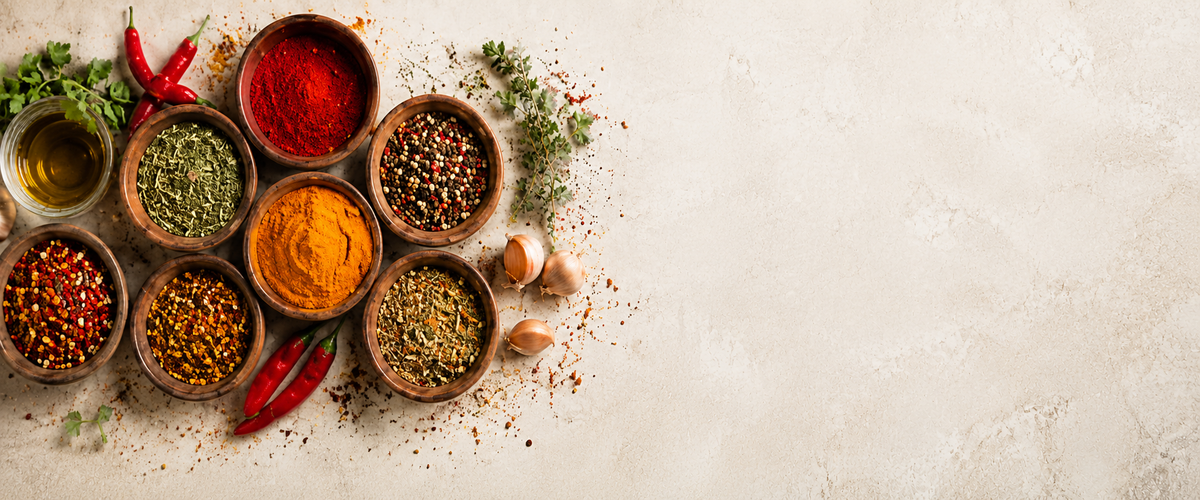 An assortment of colorful whole and ground spices in small wooden bowls, arranged on a textured light beige surface with red chili peppers, garlic cloves, and herbs. The spices include vibrant red, orange, yellow, green, and brown tones, leaving negative space on the right side.